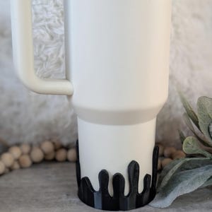 Puede incluir: Una taza de viaje blanca con un dise&ntilde;o de goteo negro alrededor de la parte inferior. La taza tiene un asa y una tapa.