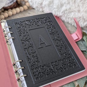 May include: A black binder with an embossed floral design and the letter "A" in the center. The binder is open, revealing white paper and a pink interior. The binder is on a wooden surface with a soft, white, fluffy background.