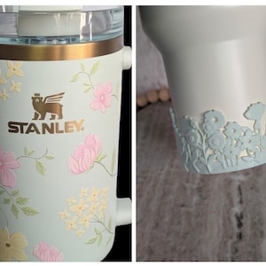 May include: A light blue Stanley tumbler with a floral design. The tumbler features pink and yellow flowers with green leaves. The Stanley logo is in gold. The bottom of the tumbler has a light blue floral cut-out design.