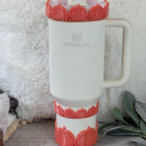 May include: White Stanley tumbler with a handle, decorated with orange pumpkin accents at the top and bottom. The tumbler has a straw and the Stanley logo. The pumpkin accents add a seasonal touch to the tumbler.