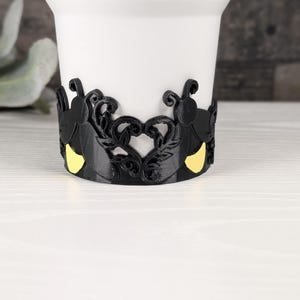 Puede incluir: Una taza blanca con una banda negra envolvente que presenta un patr&oacute;n decorativo recortado. La banda muestra siluetas de insectos negras con detalles amarillos, integradas en un dise&ntilde;o sim&eacute;trico de enredaderas y hojas en forma de coraz&oacute;n.