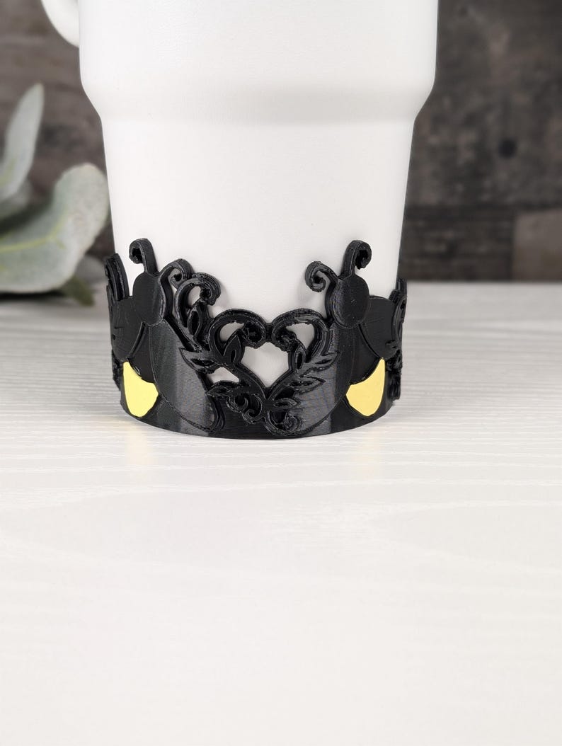 Puede incluir: Una taza blanca con una banda negra envolvente que presenta un patr&oacute;n decorativo recortado. La banda muestra siluetas de insectos negras con detalles amarillos, integradas en un dise&ntilde;o sim&eacute;trico de enredaderas y hojas en forma de coraz&oacute;n.