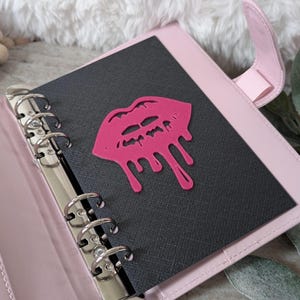 May include: A pink leather binder with a black insert featuring a hot pink lip design. The binder has silver rings and a pink strap closure. The background includes a fluffy white surface and greenery.