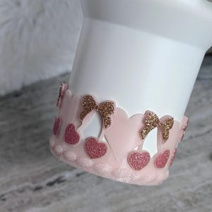 May include: A white tumbler with a pink decorative band. The band features pink hearts and gold glitter bows. The design is a mix of glitter and solid colors, creating a festive look. The tumbler is designed for beverages.