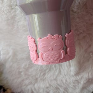 May include: A pearlescent gray tumbler with a pink owl-shaped base. The base features three pink owl designs with bows. The tumbler is designed to hold beverages and is suitable for both hot and cold drinks.