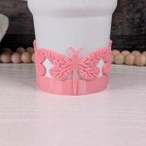 May include: A white mug featuring a pink wrap-around band at the base. The band is decorated with a repeating pattern of raised, molded dragonflies with intricate floral and leaf details on their wings, encircling the lower portion of the mug.