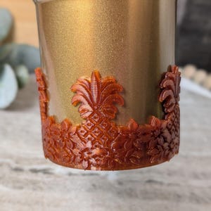 May include: A gold-colored tumbler with a detailed, reddish-brown pineapple and floral design around the base. The tumbler has a metallic finish, and the decorative elements are raised, creating a textured appearance.