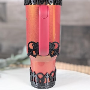 May include: A coral-colored tumbler with a glittery finish and a pink handle. The tumbler features black 3D-printed decorative bands at the top and bottom, showcasing a repeating pattern of ornate keys and heart-shaped padlocks. A clear straw is visible.