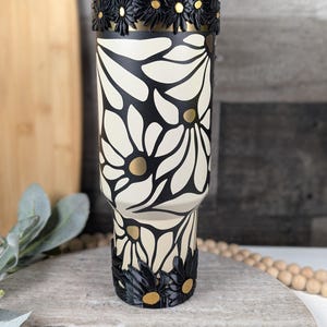 May include: A cream-colored tumbler with a black daisy floral design. The tumbler has a black rim and base with daisy accents and gold centers. The design is repeated around the body of the tumbler. The tumbler is approximately 10 inches tall.