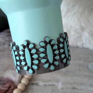 May include: A light teal tumbler with a decorative band of gray and teal floral accents. The band is made of a series of teardrop-shaped elements, each with a teal center. The tumbler has a smooth, matte finish.