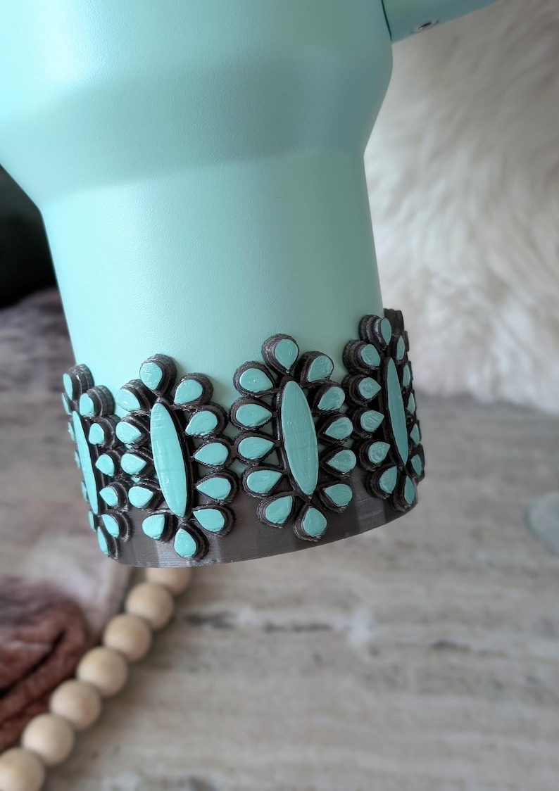 May include: A light teal tumbler with a decorative band of gray and teal floral accents. The band is made of a series of teardrop-shaped elements, each with a teal center. The tumbler has a smooth, matte finish.