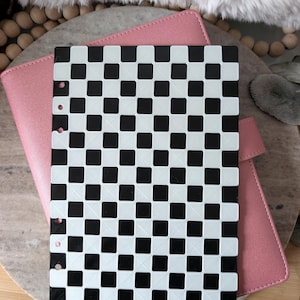 May include: A black and white checkered planner insert is placed over a pink planner. The insert has several holes for binding. The planner is on a wooden surface.