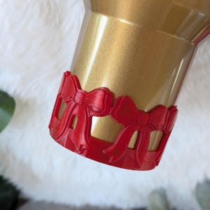 May include: Gold tumbler with a red bow-shaped trim around the bottom.