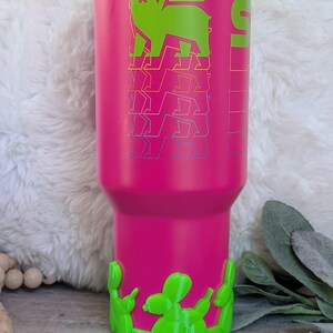 May include: A vibrant pink tumbler with a green bear graphic and matching green base. The bear has wings and a crown. The tumbler is designed to keep drinks cold or hot. A stylish and functional drinkware item.