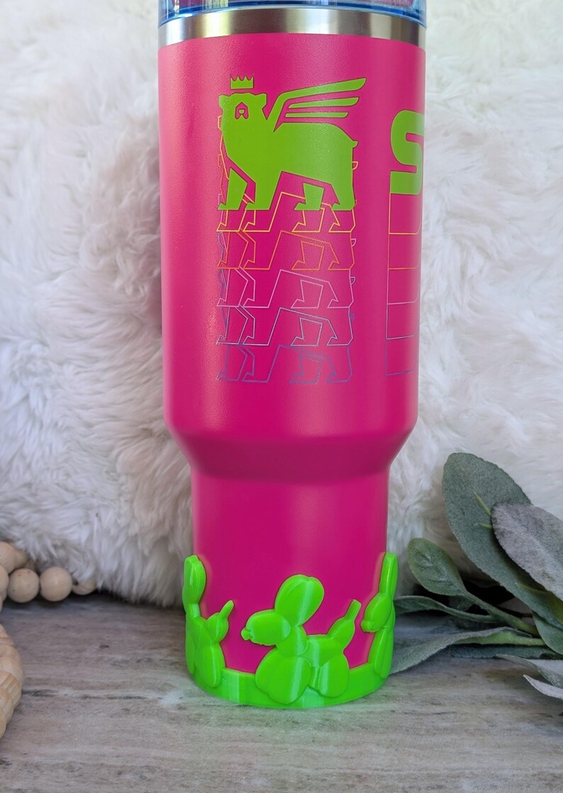 May include: A vibrant pink tumbler with a green bear graphic and matching green base. The bear has wings and a crown. The tumbler is designed to keep drinks cold or hot. A stylish and functional drinkware item.