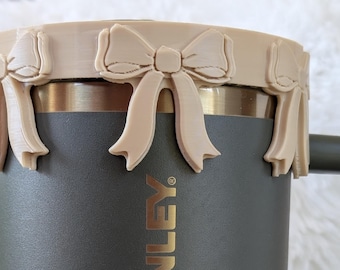 May include: A gray stainless steel travel mug with a black handle. The mug has a beige silicone lid with a decorative bow design.