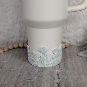May include: A white tumbler with a floral design around the base. The tumbler has a handle and a smooth, matte finish. The floral design is a lighter shade of white, creating a subtle contrast.
