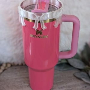 May include: A pink Stanley travel mug with a white bow and a clear straw. The mug has a handle and a gold logo that says "STANLEY".
