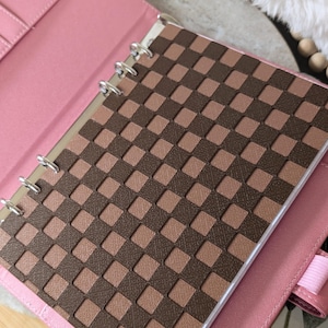 May include: Pink binder with a brown and tan woven design on the front cover. The binder is open, revealing the interior pages and silver metal rings. The cover has a pink leather-like texture.