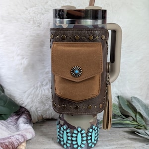 May include: A camouflage-patterned tumbler with a brown leather wrap featuring a small pocket and a turquoise-centered decorative button. The bottom of the tumbler has a turquoise floral design. A tan handle and a brown straw complete the design.