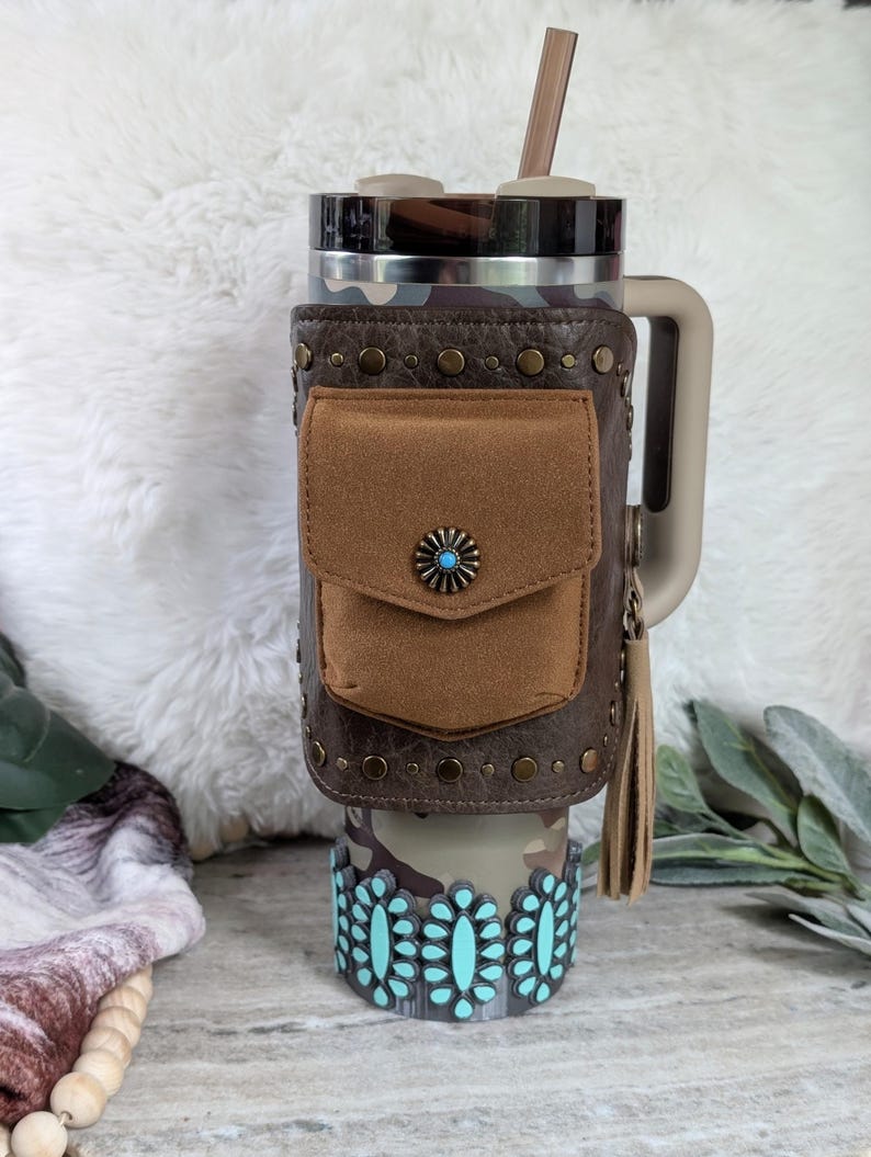 May include: A camouflage-patterned tumbler with a brown leather wrap featuring a small pocket and a turquoise-centered decorative button. The bottom of the tumbler has a turquoise floral design. A tan handle and a brown straw complete the design.