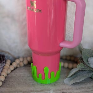 May include: A bright pink Stanley tumbler with a matching pink handle. The tumbler has a gold rim and the Stanley logo in gold. A green, drippy-style accessory is at the base of the tumbler.