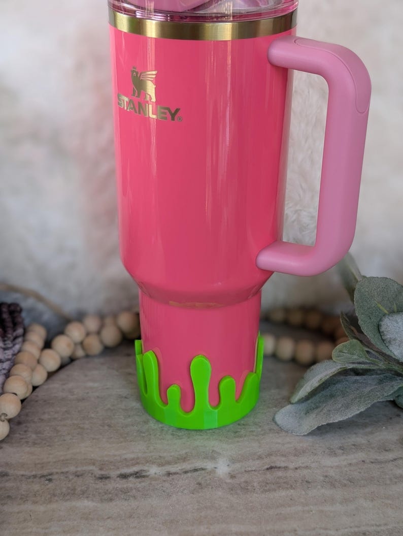 May include: A bright pink Stanley tumbler with a matching pink handle. The tumbler has a gold rim and the Stanley logo in gold. A green, drippy-style accessory is at the base of the tumbler.