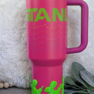 May include: A pink insulated tumbler with a handle and a straw. The word "STANLEY" is printed in green with a wavy design. The base features green balloon dog accents. The lid is blue and purple.
