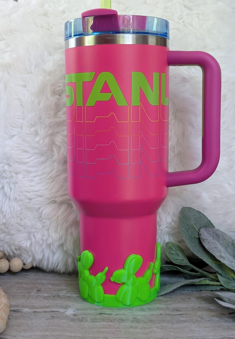 May include: A pink insulated tumbler with a handle and a straw. The word "STANLEY" is printed in green with a wavy design. The base features green balloon dog accents. The lid is blue and purple.