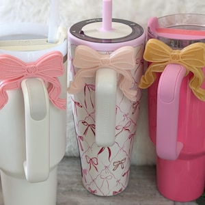 May include: Three insulated tumblers with handles, each adorned with a decorative bow. One tumbler is white with a pink bow, another is pink with a yellow bow, and the third is patterned with pink bows.