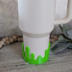 May include: A white Stanley tumbler with a bright green drip-style base. The tumbler has a handle and the Stanley logo is visible. The green base adds a playful, decorative element to the tumbler.