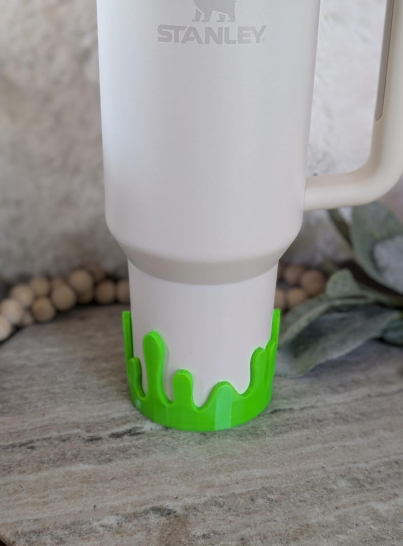 May include: A white Stanley tumbler with a bright green drip-style base. The tumbler has a handle and the Stanley logo is visible. The green base adds a playful, decorative element to the tumbler.