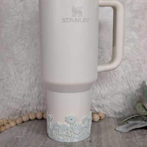 May include: A white Stanley travel mug with a light gray lid and handle. The Stanley logo is visible on the front. The bottom of the mug features a floral design. The mug is designed to hold a large quantity of liquid.