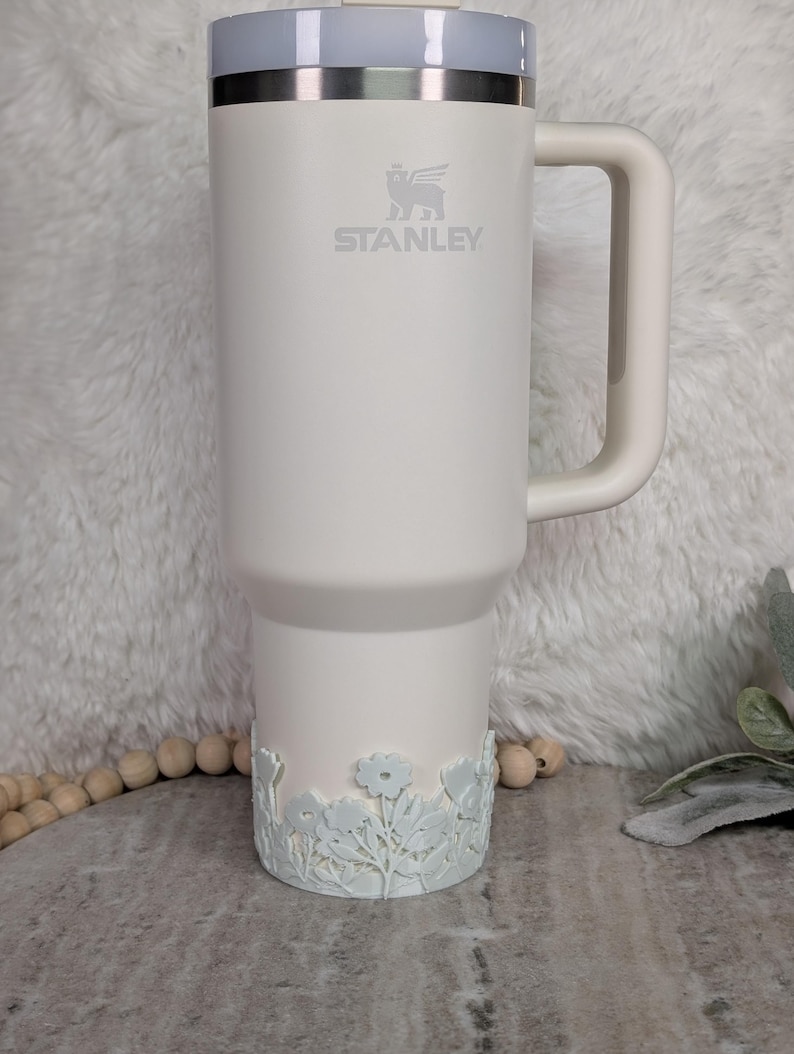 May include: A white Stanley travel mug with a light gray lid and handle. The Stanley logo is visible on the front. The bottom of the mug features a floral design. The mug is designed to hold a large quantity of liquid.