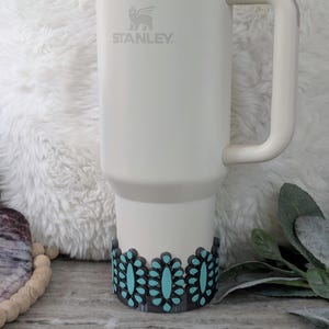May include: A white Stanley tumbler with a handle, featuring a turquoise and black floral design around the base. The Stanley logo is visible on the front. The tumbler is designed for beverages.