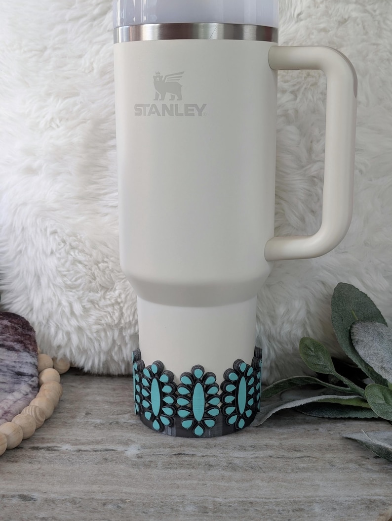 May include: A white Stanley tumbler with a handle, featuring a turquoise and black floral design around the base. The Stanley logo is visible on the front. The tumbler is designed for beverages.
