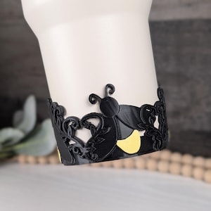 Puede incluir: Una banda decorativa negra envuelta alrededor de un vaso blanco, con un patr&oacute;n repetitivo de insectos estilizados con cuerpos negros y detalles amarillos, intercalados con intrincados recortes de hojas y enredaderas.
