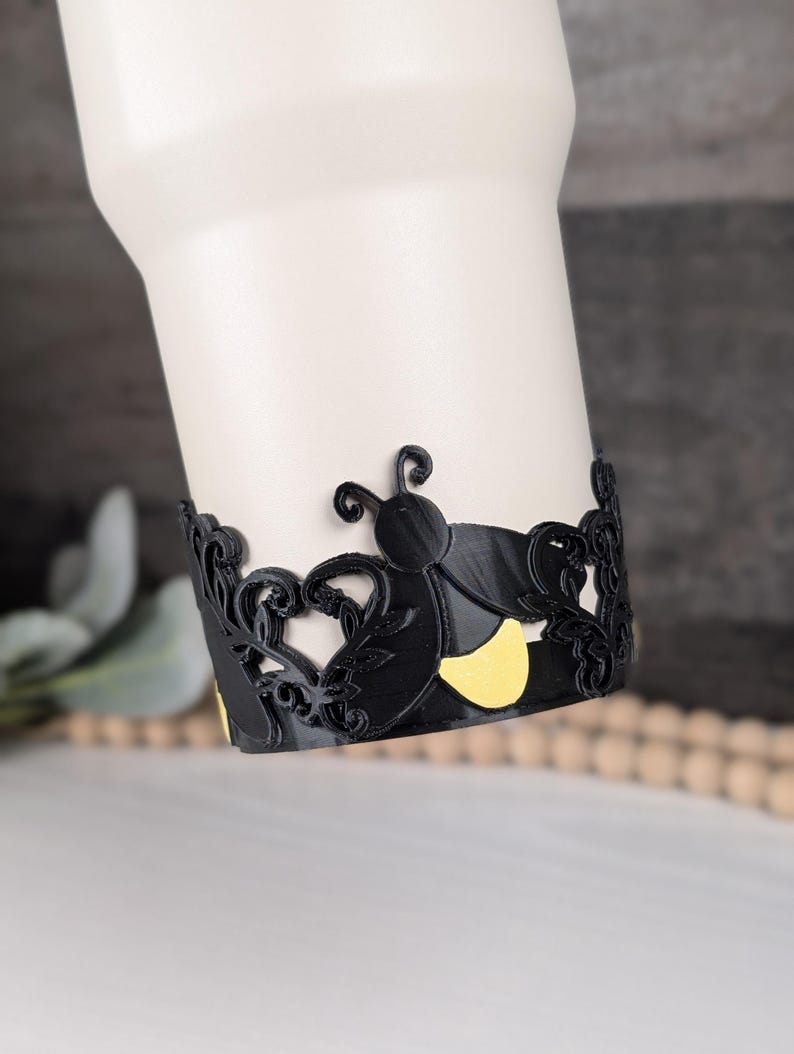 Puede incluir: Una banda decorativa negra envuelta alrededor de un vaso blanco, con un patr&oacute;n repetitivo de insectos estilizados con cuerpos negros y detalles amarillos, intercalados con intrincados recortes de hojas y enredaderas.