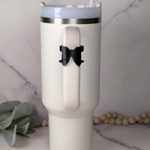 Puede incluir: Un vaso térmico de color blanco mate con un asa resistente que presenta un pequeño lazo negro tridimensional sujeto en el centro. El vaso incluye una pajita de plástico transparente y una tapa blanca con un borde de metal plateado.