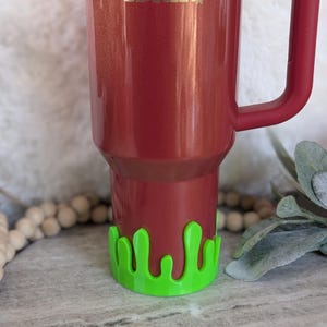 May include: A red Stanley tumbler with a green slime-shaped base. The tumbler has a handle and the Stanley logo in gold. The base is a bright green color and adds a fun, unique touch to the tumbler.