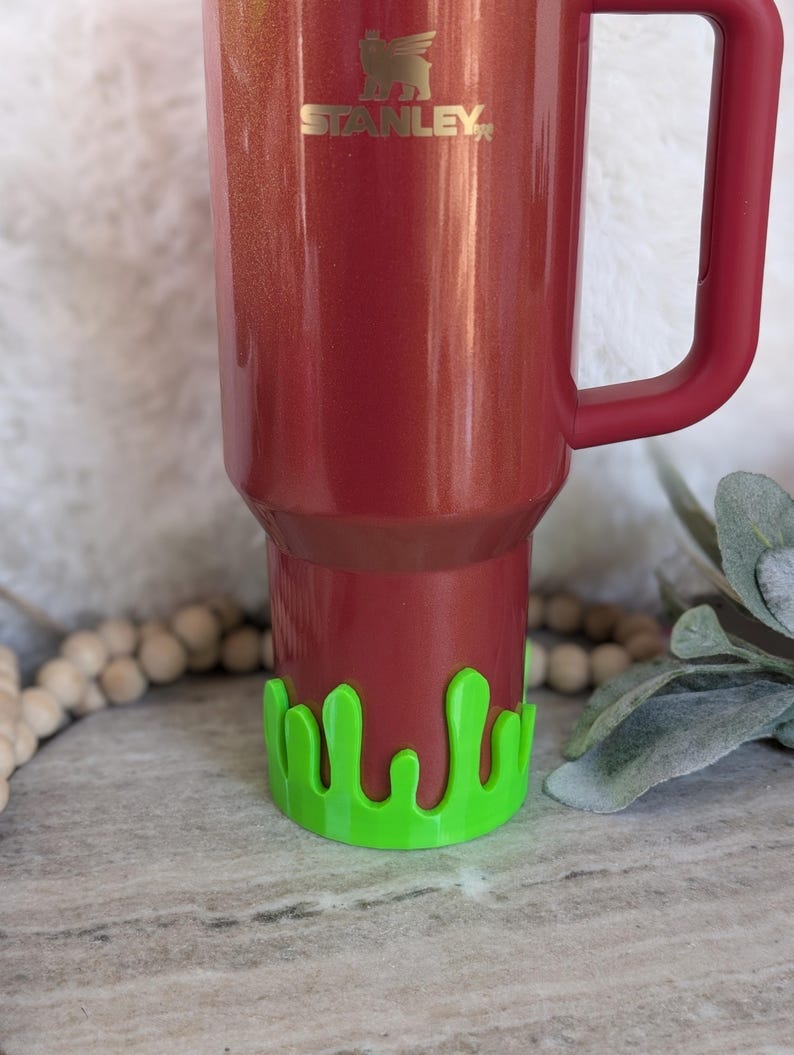 May include: A red Stanley tumbler with a green slime-shaped base. The tumbler has a handle and the Stanley logo in gold. The base is a bright green color and adds a fun, unique touch to the tumbler.