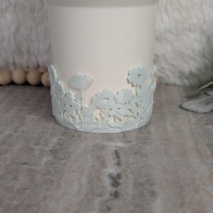 May include: A white ceramic candle holder with a floral design around the base. The flowers are a light blue color and are raised from the surface. The candle holder is cylindrical and has a smooth finish.