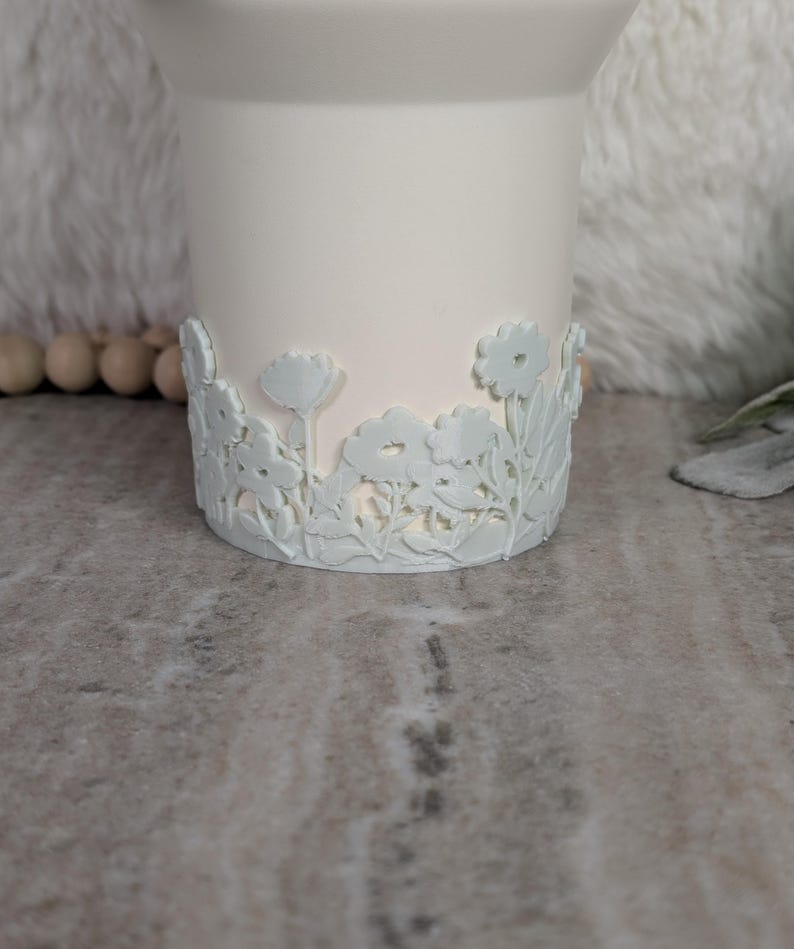 May include: A white ceramic candle holder with a floral design around the base. The flowers are a light blue color and are raised from the surface. The candle holder is cylindrical and has a smooth finish.