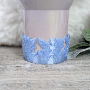 May include: A light blue, iridescent tumbler with a decorative, 3D printed, light blue fairy design around the bottom.