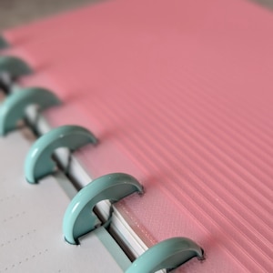 May include: Close-up of a planner with a pink cover and lined pages. The planner is held together by light teal metal rings. The cover has a textured, ribbed design. The pages are white with dotted lines.