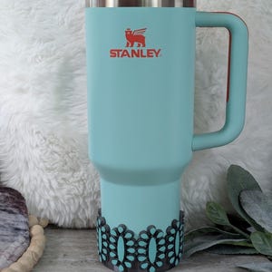 May include: A teal Stanley tumbler with a red lid and handle. The tumbler has the Stanley logo in red and a decorative black and teal base. The tumbler is designed for cold beverages and has a straw.