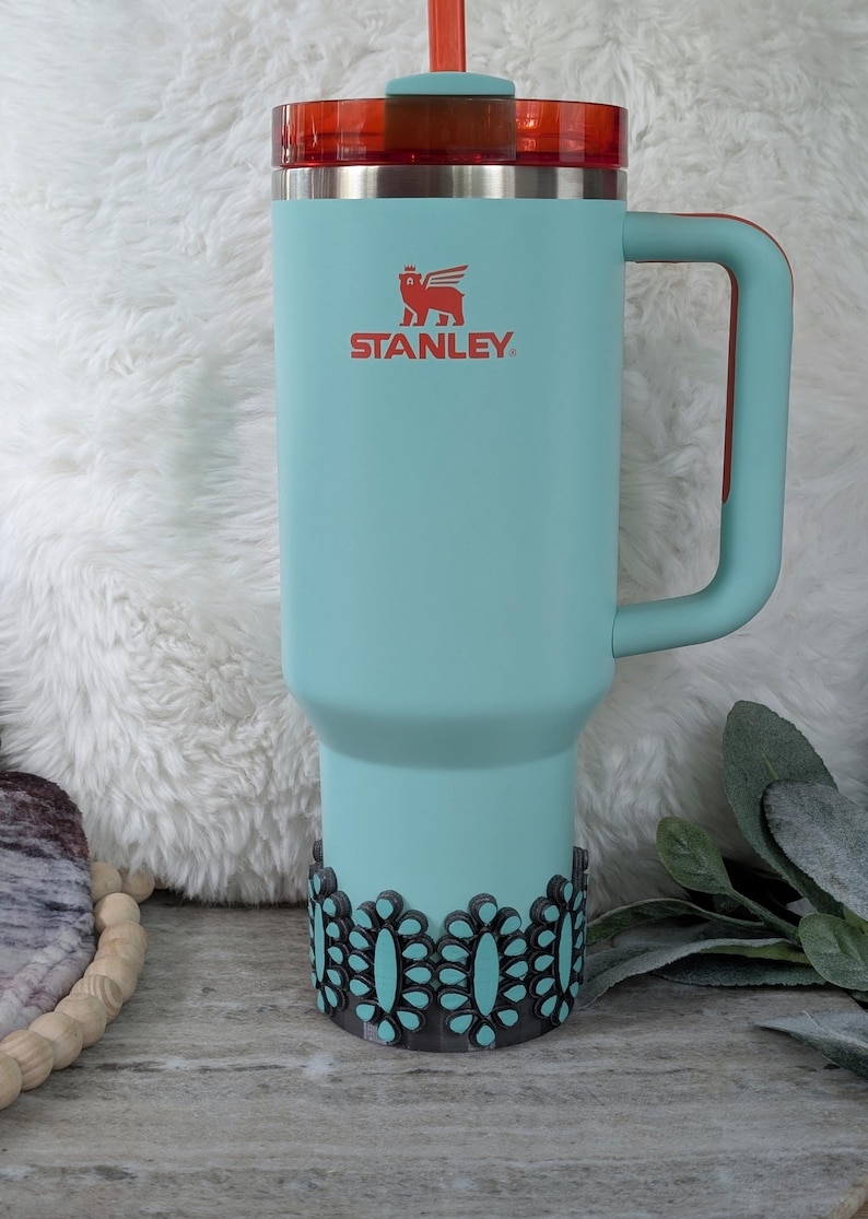 May include: A teal Stanley tumbler with a red lid and handle. The tumbler has the Stanley logo in red and a decorative black and teal base. The tumbler is designed for cold beverages and has a straw.