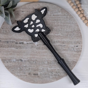 May include: A black and white cow-shaped bottle stopper. The cow head has white spots and is decorated with black roses. The stopper has a long, textured black handle. This decorative item is designed for wine bottles.