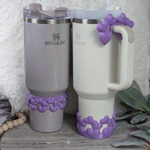 May include: Two Stanley tumblers, one gray and one white, with clear lids and straws. Both tumblers have a purple heart-shaped embellishment around the base. The gray tumbler has a matching heart embellishment on the lid. The Stanley logo is visible on both tumblers.