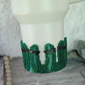 May include: White tumbler with a green, 3D-printed base featuring cartoon pickles wearing sunglasses. The pickles are a vibrant green, and the sunglasses are black. The tumbler has a handle and a smooth, matte finish.